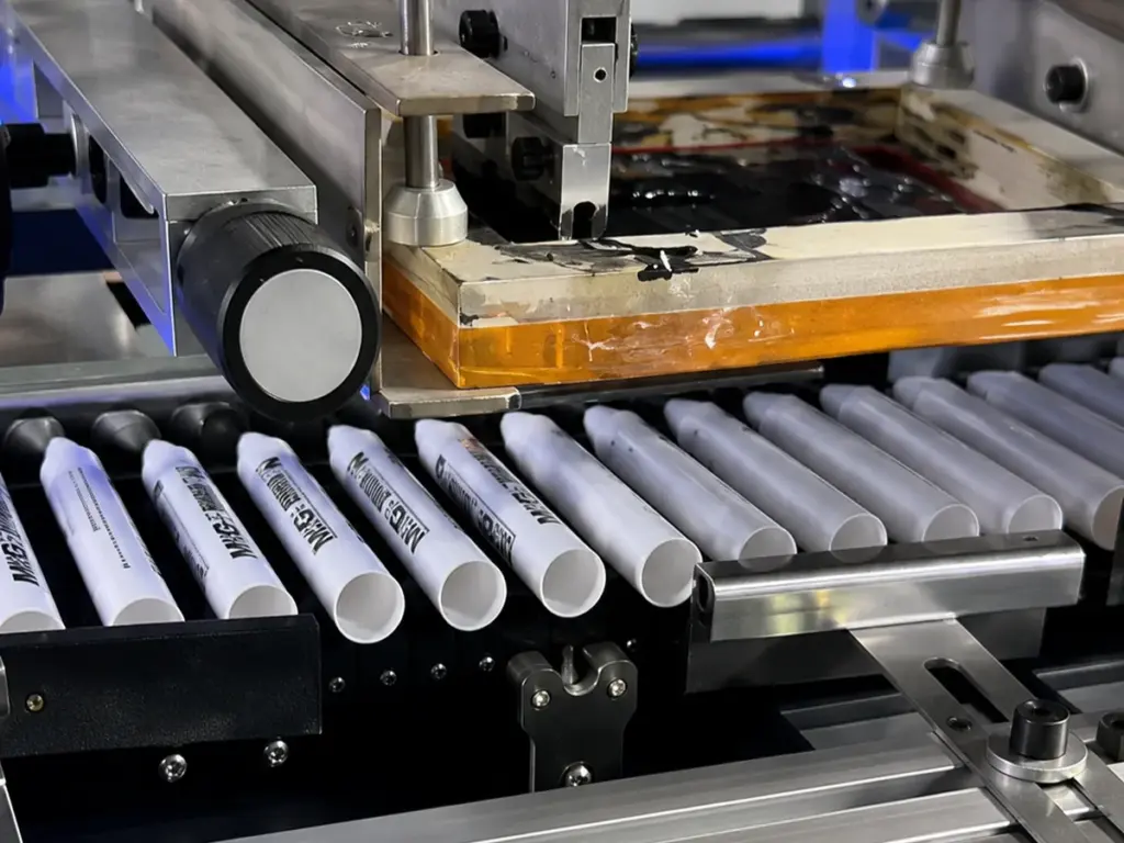 Automated screen printing machine processing white pen barrels on conveyor.