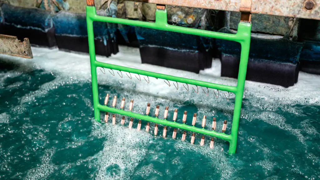 Metal pen components on a green rack undergoing electroplating in a bubbling industrial chemical bath.
