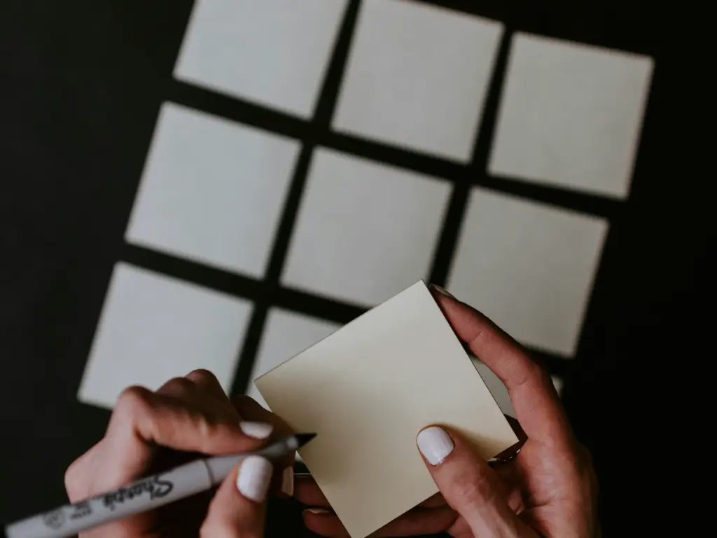 A pair of hands writing on a sticky note with a grid of nine blank notes arranged on a dark surface.