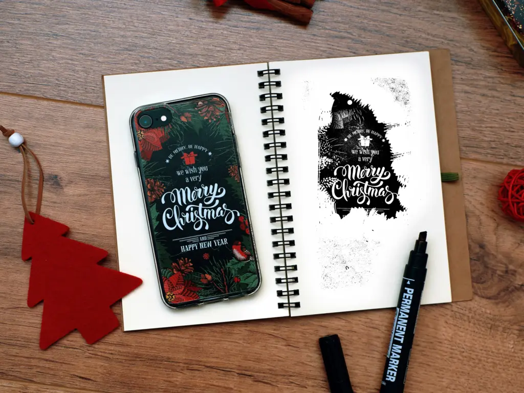 Open sketchbook with Merry Christmas calligraphy and a matching festive phone case on a wooden desk