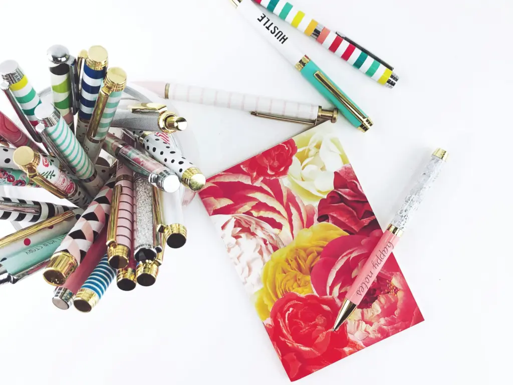 Collection of colorful patterned pens in a cup alongside a floral note card and decorative pens on a white surface
