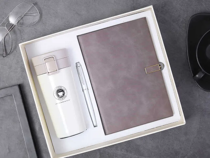 Top view of a gift box containing a brown notebook, white travel mug, and silver pen on a grey surface