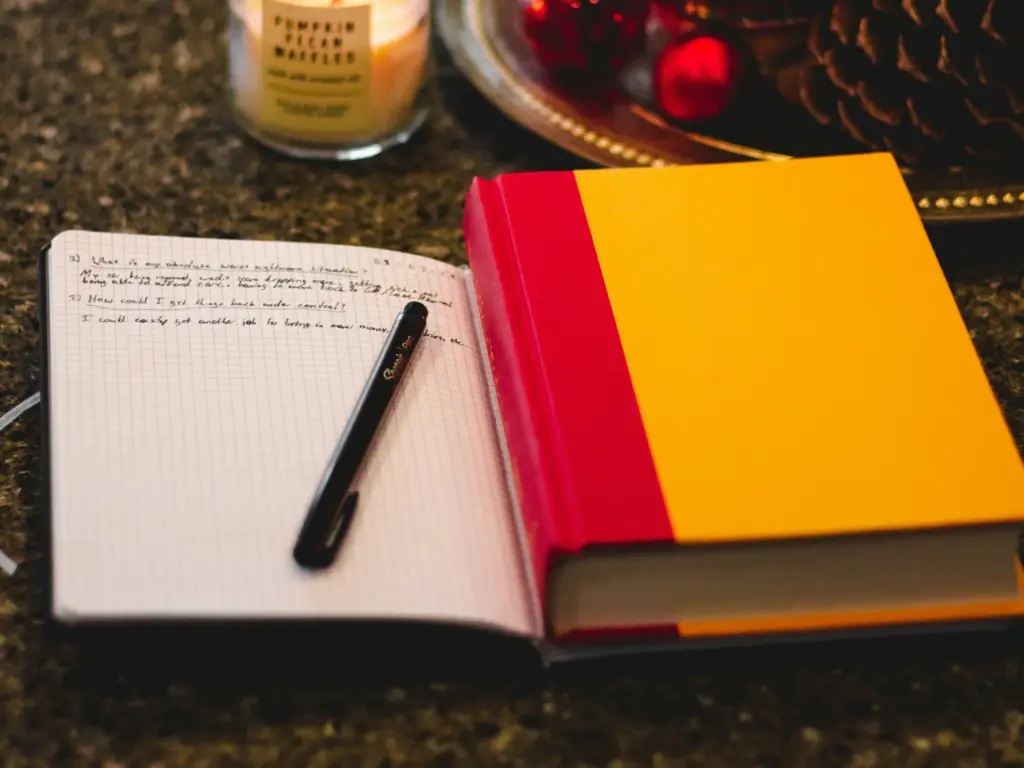 Open journal with handwritten notes and pen beside a lit candle and Christmas ornaments on a granite countertop