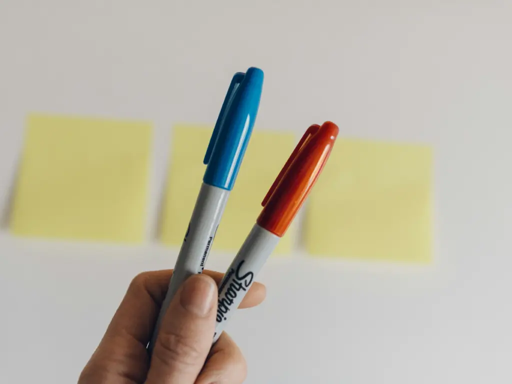 A hand holding two Sharpie markers with blue and orange caps against yellow sticky notes